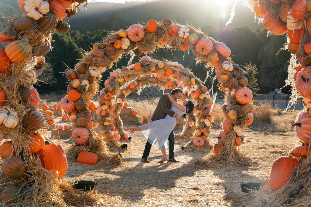 Engaged indian couple under a pumpkin arch the woman is wearing a light blue summer dress and is being dipped by her fiancé with her one leg sticking out smiling at one another