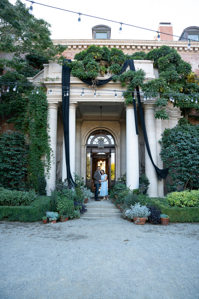 Indian couple standing in front of the Filoli Gardens Museum entrance staring into each other's eyes. The woman is dressed in a light blue dress and the man is wearing a grown blazer and dark blue washed jeans