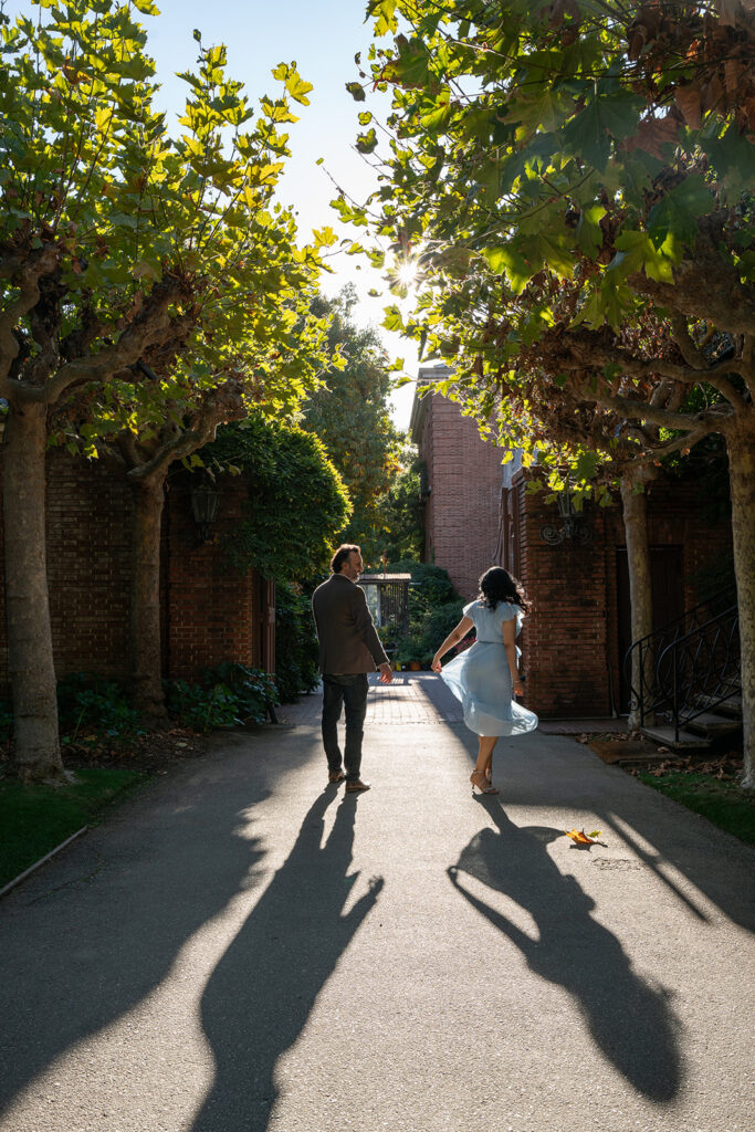 Indian woman in her light blue dress is dancing next to her fiancé who is dressed in a brown blazer and blue jeans and their shadows follow behind them imitating their movements