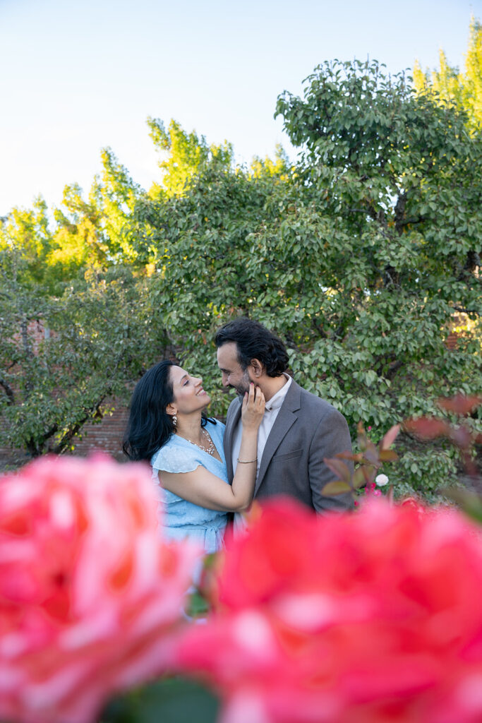 Indian woman in her blue dress is reaching up touching her fiancé's face who is smiling down at her looking at her lovingly as the pink flowers outline the bottom frame