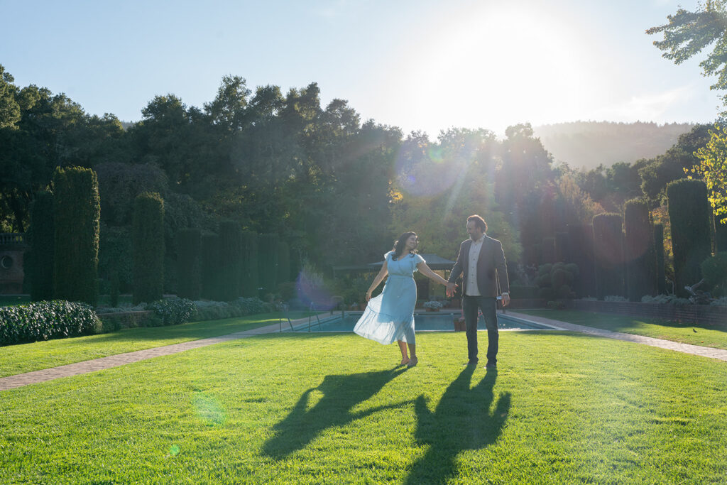 Indian couple holding hands and dancing in an open grass field with their shadows dancing along with them during high noon and they are both smiling at one another