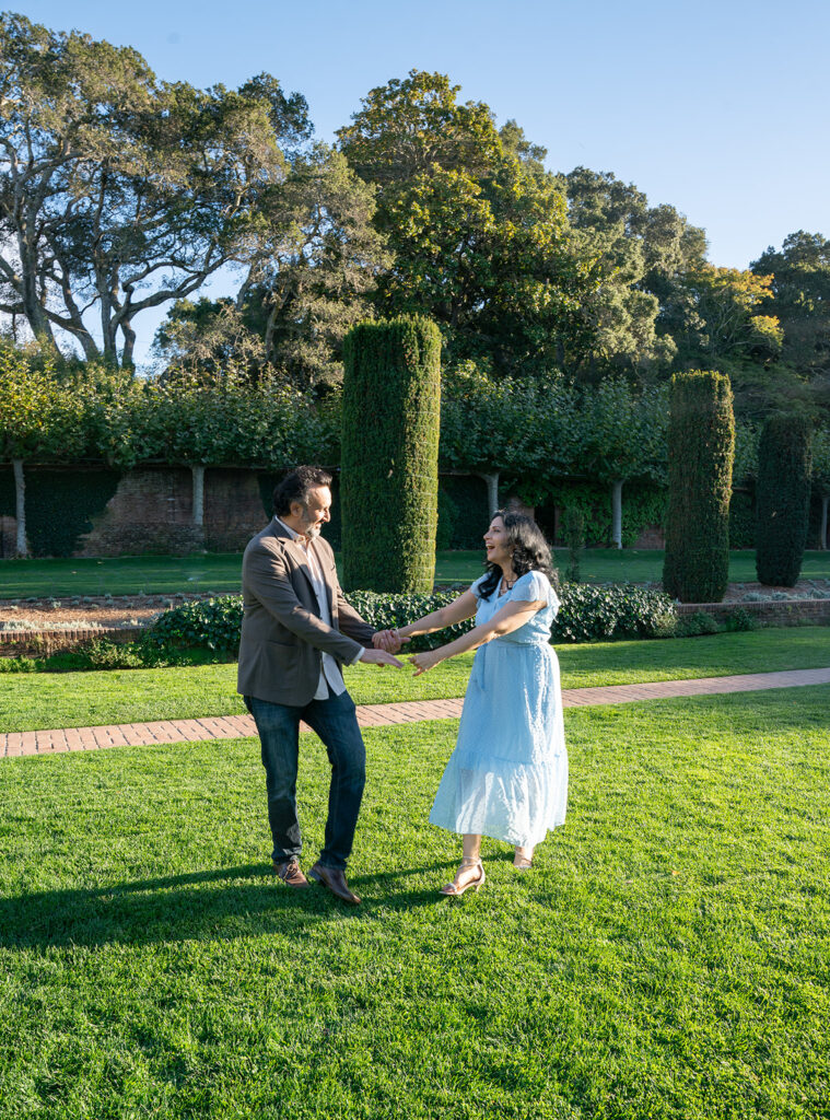 Indian couple holding hands and dancing in an open grass field with their shadows dancing along with them during high noon and they are both smiling at one another