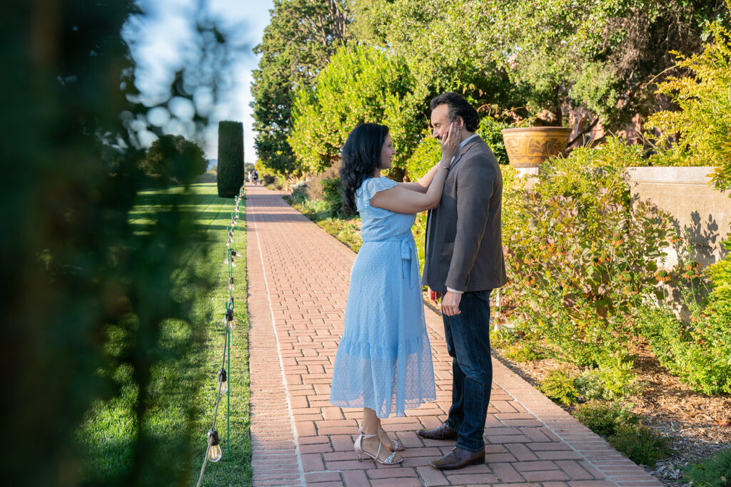 Photo of Indian couple looking up and down at one another and the woman in the light blue dress is gently touching his face while the edge of a hedge frames the left side of them