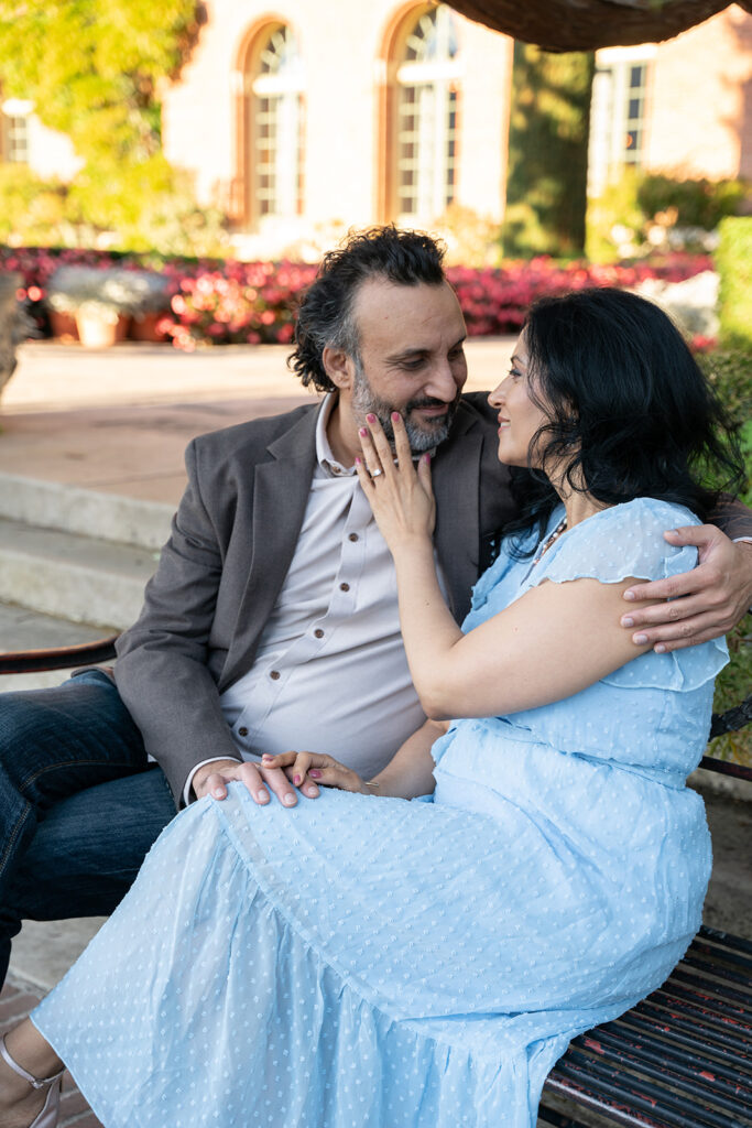 Indian man dressed in brown blazer and beige button down holds his fiance who is jin a light blue dress on a bench staring at her while she gently touches her face