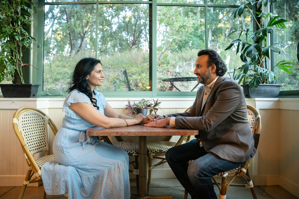 Indian couple sitting on oppositen sides of a coffee table at a cafe holding hands across the table smiling with a bright window behind them with trees and bushes behind them