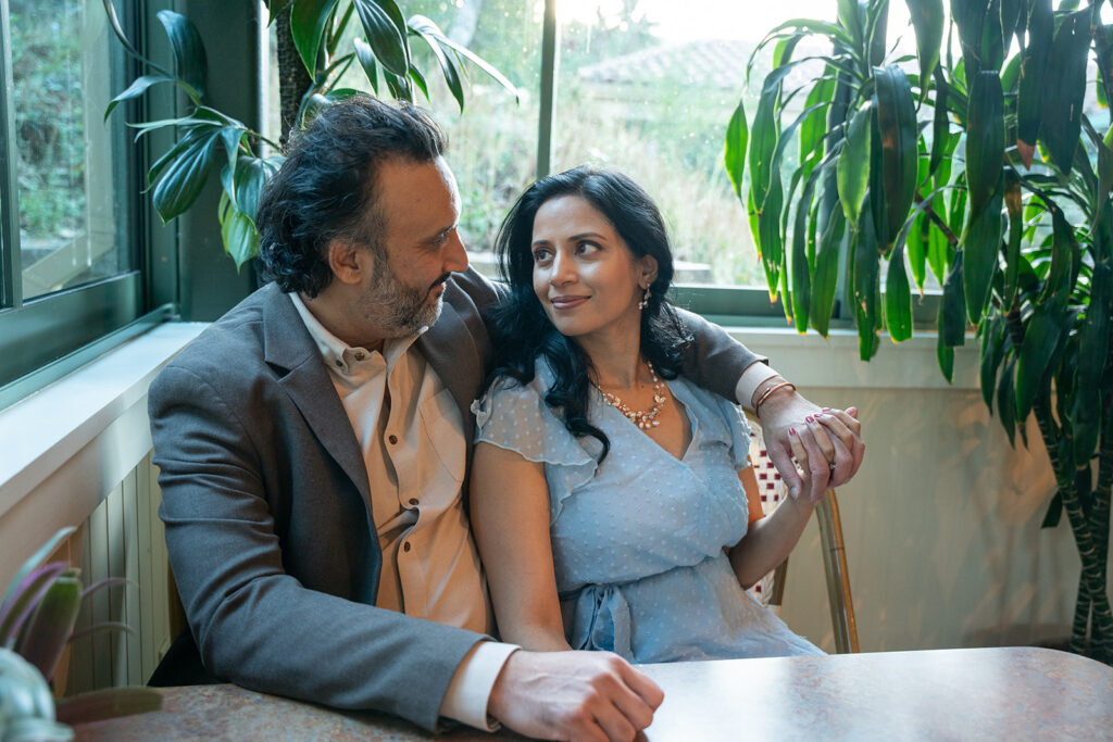 Indian couple sitting side by side in a cafe with the woman in a blue dress gently lays on his left shoulder staring up into his eyes while the man is wearing a brown blazer and beige top staring down at her smiling back