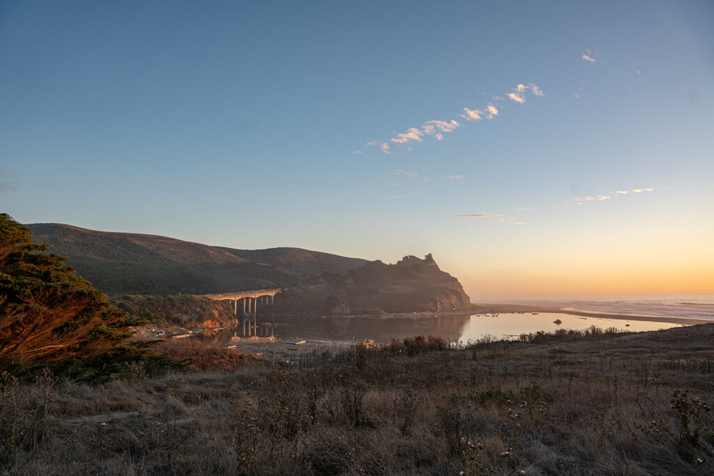 Mountain landscape shot at sunset of a bridge connected to mountains and the ocean underneath it with a couple of clouds going diagonally upright