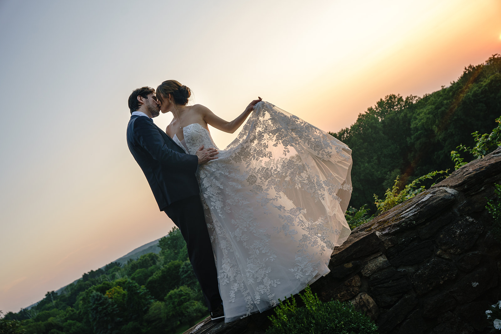 Wedding couple standing on a stone wall at sunset kissing each other in South Salem , New York at Le Chateau