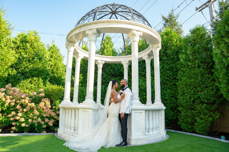 Beautiful black wedding couple pose in front of a white gazebo surrounded greenery and flowers