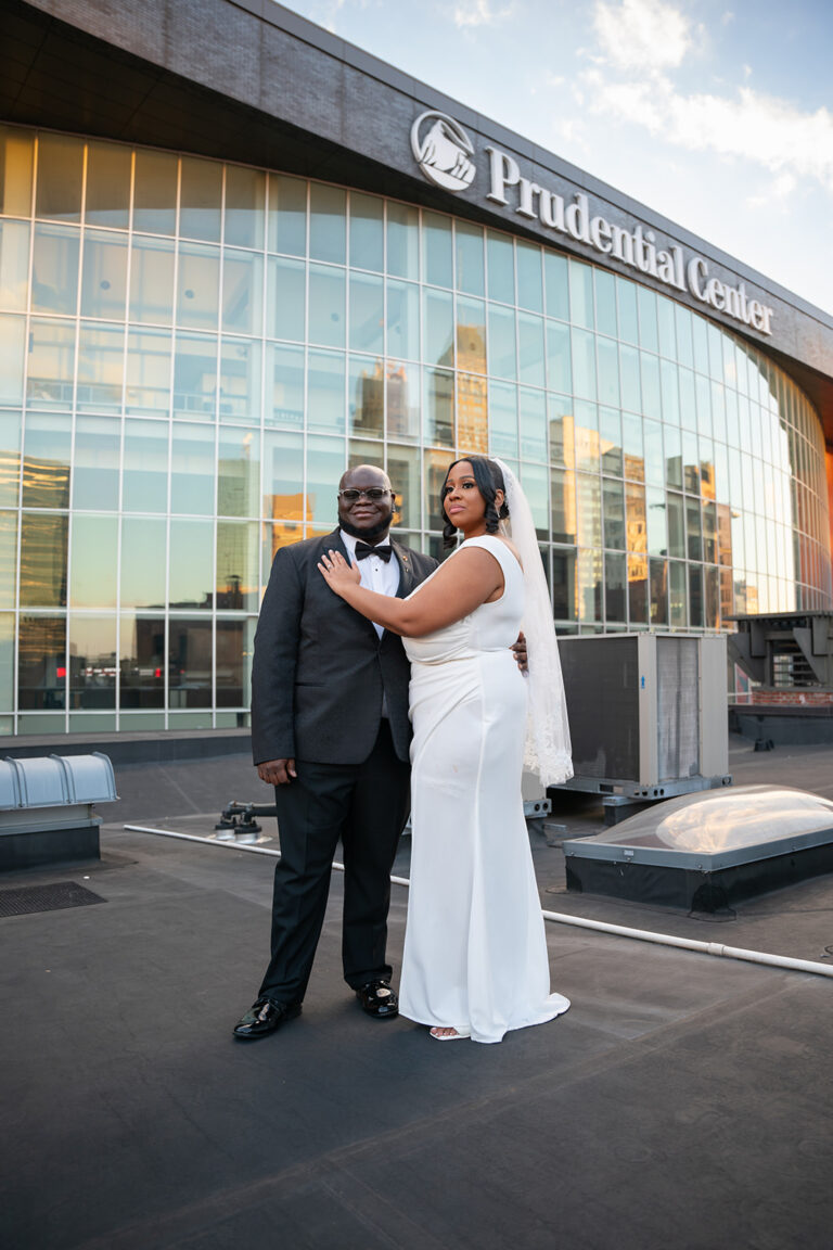Black couple posing on their wedding day on a rooftop with Newark Prudential center in the background in Newark, New Jersey