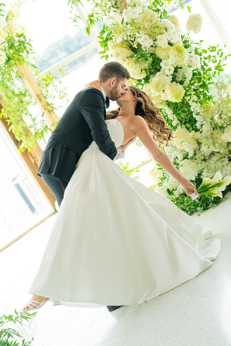 Jewish bride being dipped under the Huppah by her groom eyes closed kissing while they are framed by bright sunlight and floral decorations at The Lighthouse venue at Chelsea Piers in New York City.